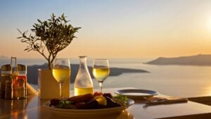 This image shows a table set for two at sunset in Santorini. There's a carafe and two glasses of white wine, and a platter with Greek food.