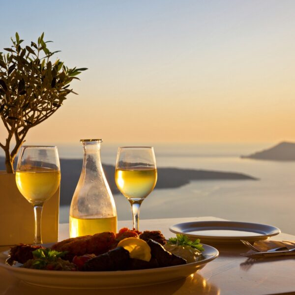 This image shows a table set for two at sunset in Santorini. There's a carafe and two glasses of white wine, and a platter with Greek food.