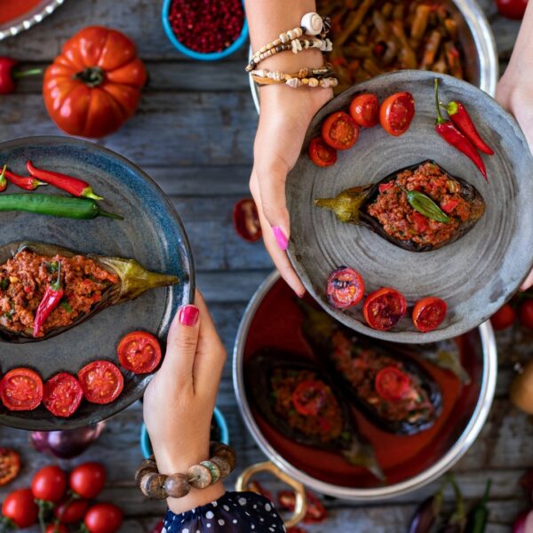 This is a close-up of the hands of two women holding one dish of imam bayildi (stuffed eggplant) each.