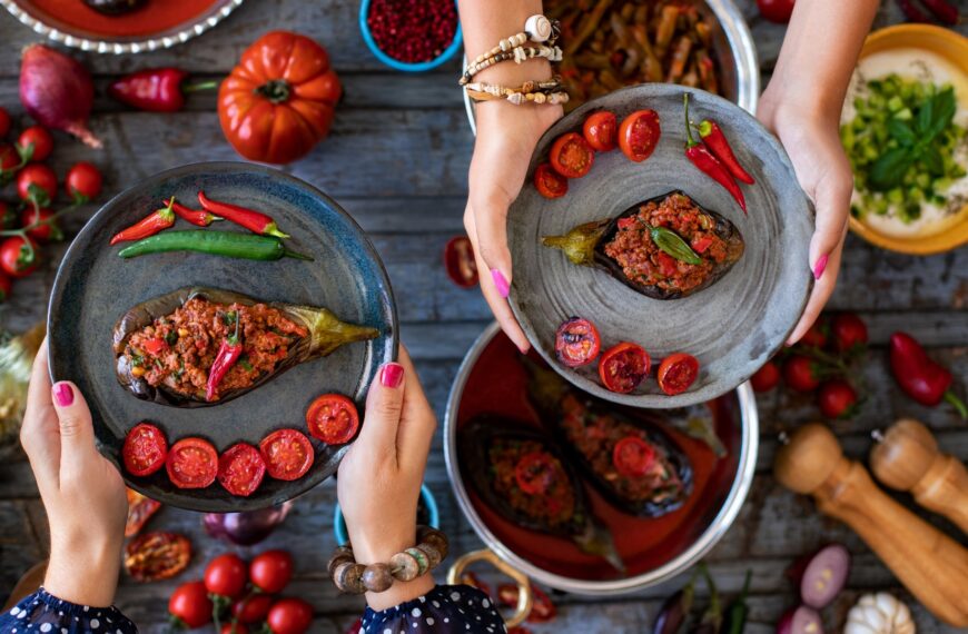 This is a close-up of the hands of two women holding one dish of imam bayildi (stuffed eggplant) each.