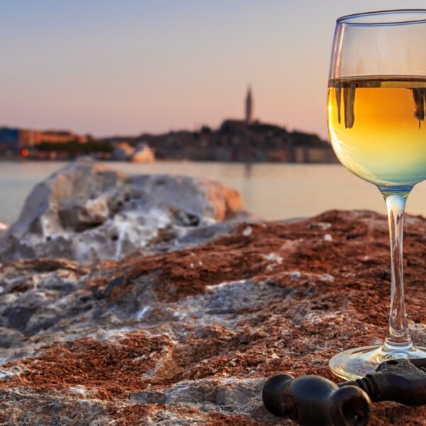 This image shows a glass of white wine on a rock with the town of Rovinj in the background. This is the featured image of our guide to the best wineries in Istria, Croatia.