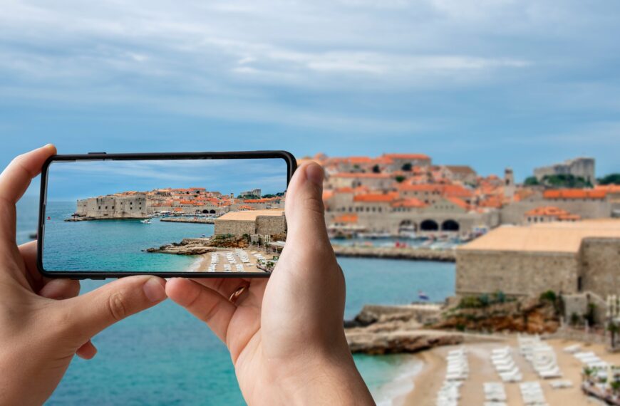 This is a panoramic view of Banje Beach with its turquoise waters and golden sand. In the background, the walled Old Town of Dubrovnik with the iconic red rooftops spreads in all its glory. In the foreground, two hands holding a phone, taking a picture of the landscape.