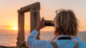 This image shows a woman taking a photo of Portara at sunset with her mobile phone.